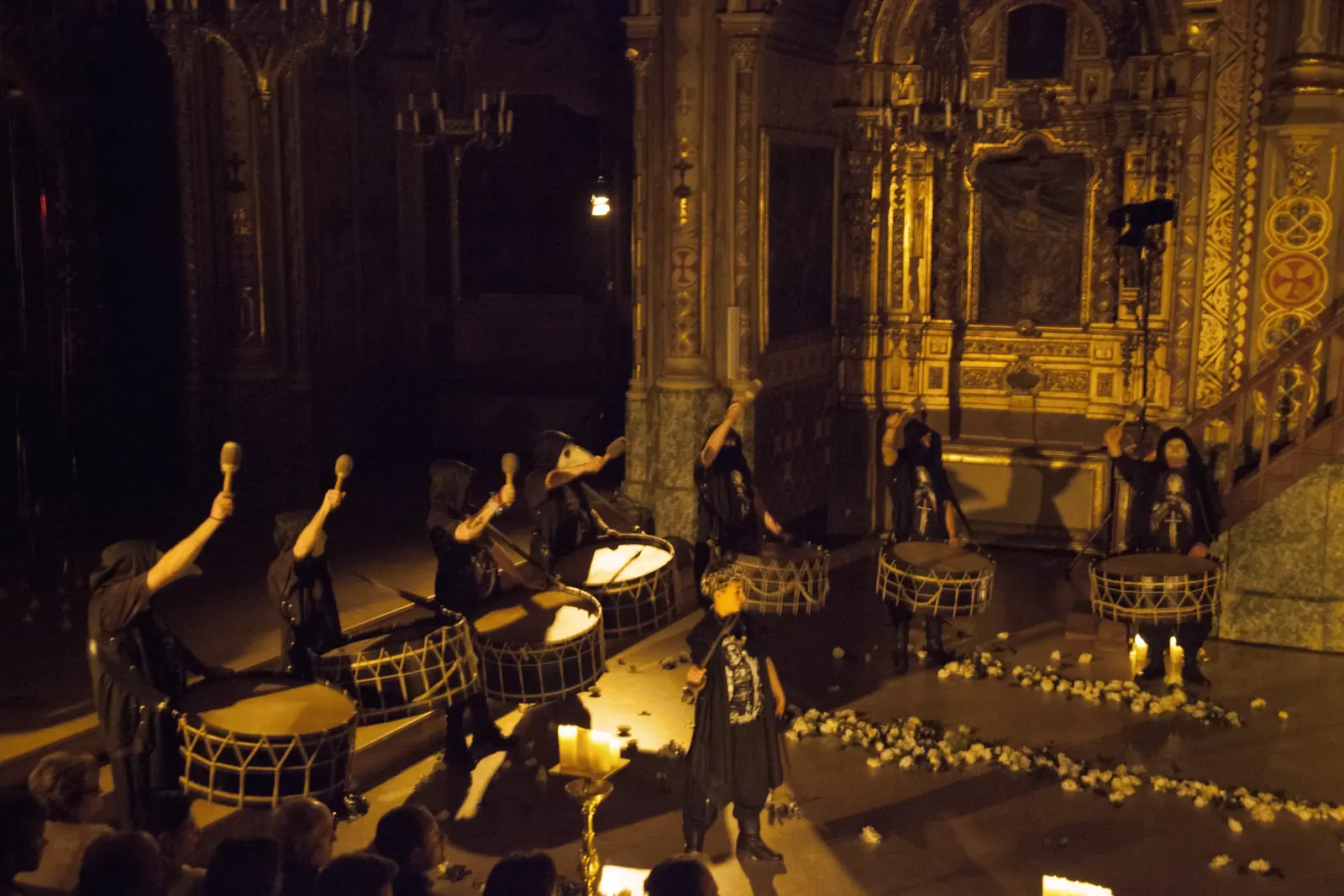 Hooded drummers striking the bass drums in the candlelit nave