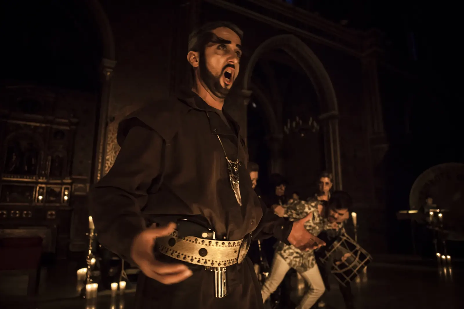 A bearded singer in a dark tunic mid-aria, dancers behind him