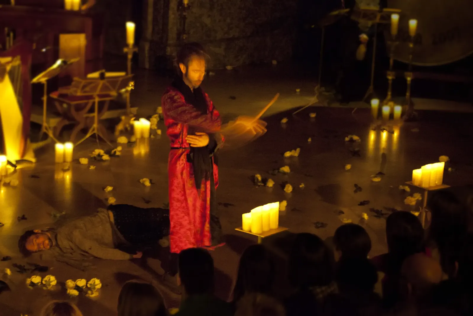 A figure in red standing over a fallen man, candles all around